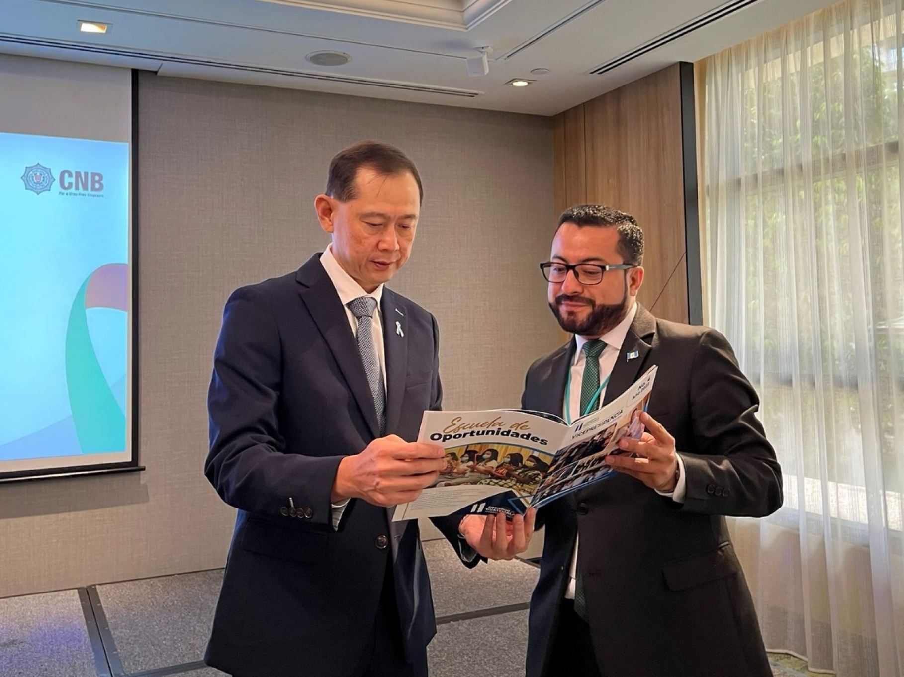 Two men in suits read a brochure titled "Escuela de Oportunidades". A CNB logo is displayed on a screen in the background.